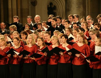 Foto: Der Universitätschor steht in einer Kirche und singt. Die Chormitglieder sind in rot bzw. schwarz gekleidet und halten ihre Singebücher in der Hand