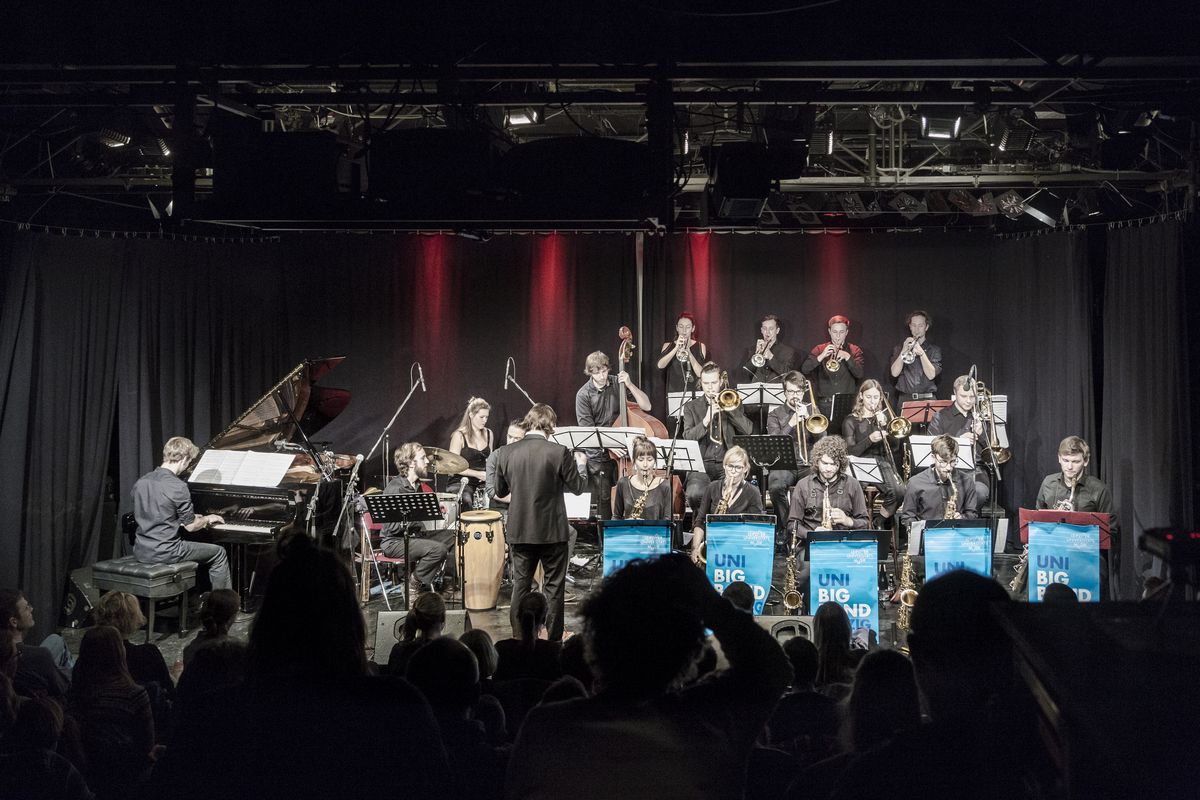 zur Vergrößerungsansicht des Bildes: Aufnahme von zahlreichen Musikern mit Blasinstrumenten sowie einem Klavier auf einer Bühne
